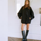 A woman stands in a bright white room with a wood herringbone floor, wearing an oversized black coat, black shorts, and black knee-high boots.