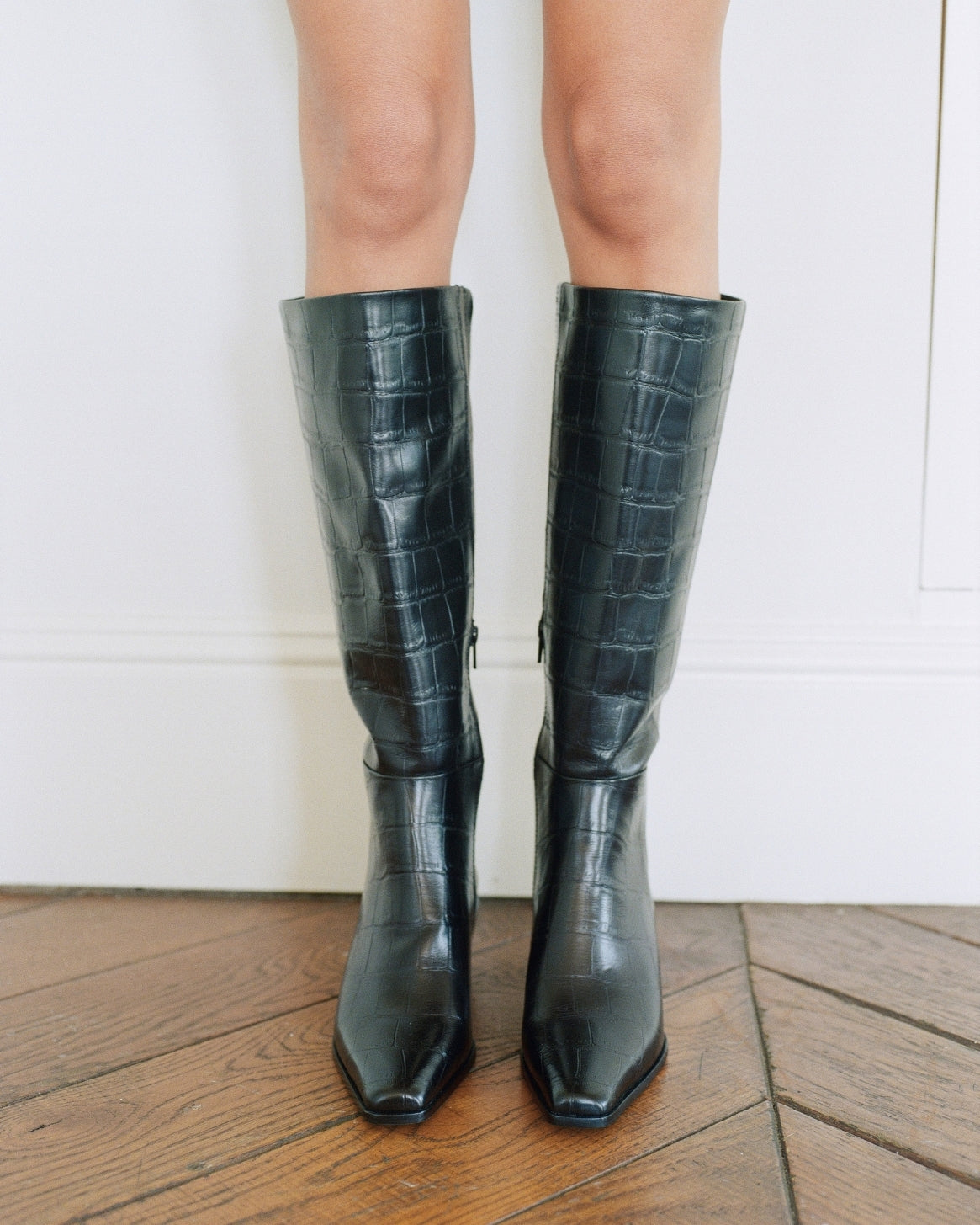 A person wearing knee-high black leather boots with an embossed crocodile pattern, standing on a wooden floor against a white wall.