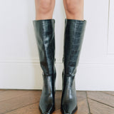 A person wearing knee-high black leather boots with an embossed crocodile pattern, standing on a wooden floor against a white wall.
