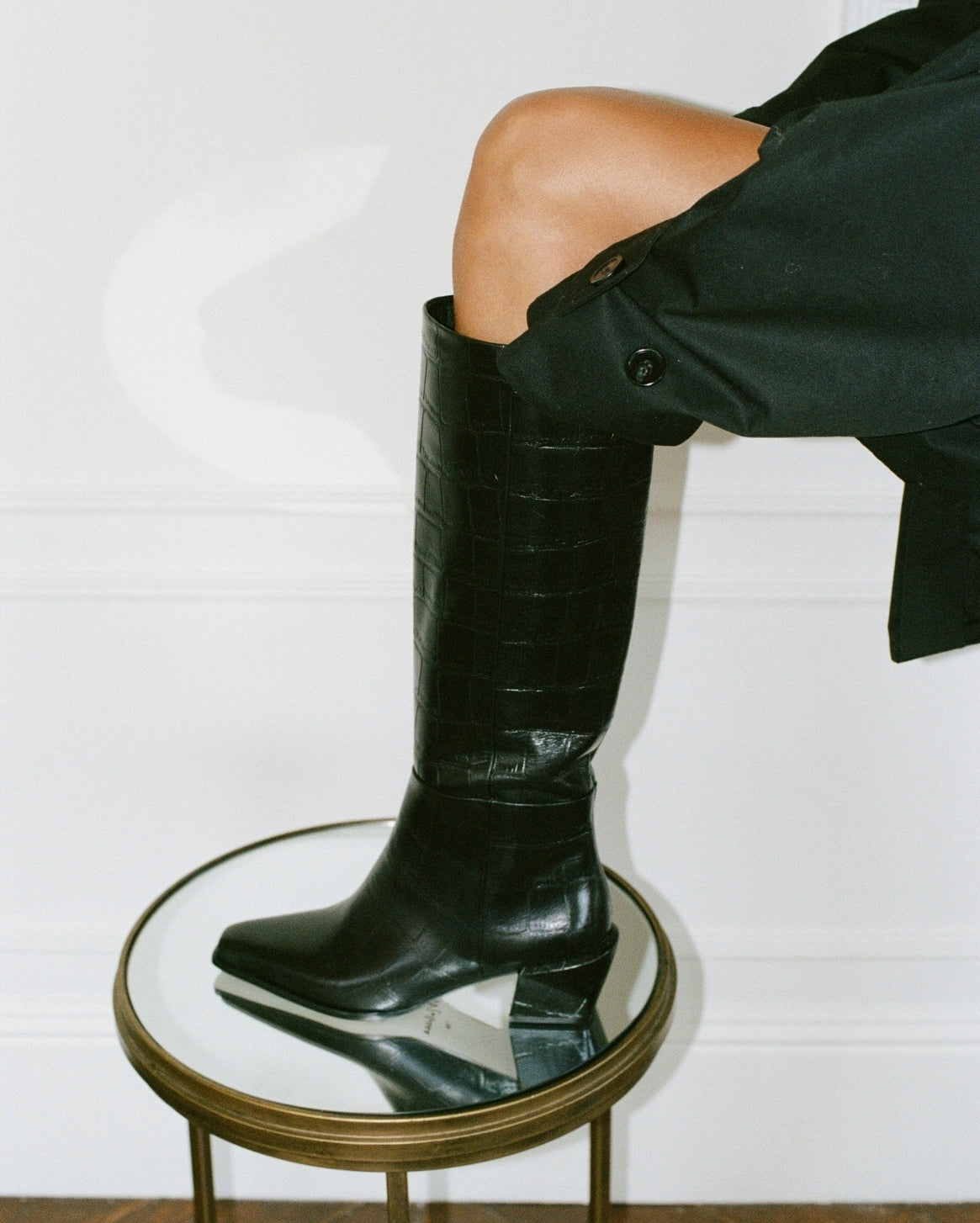A black crocodile-embossed leather knee-high boot with a chunky block heel, worn on a model's leg who is also wearing a black coat. The boot is styled on a small round mirrored stool with a gold frame against a white paneled wall.