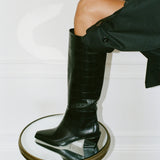 A black crocodile-embossed leather knee-high boot with a chunky block heel, worn on a model's leg who is also wearing a black coat. The boot is styled on a small round mirrored stool with a gold frame against a white paneled wall.
