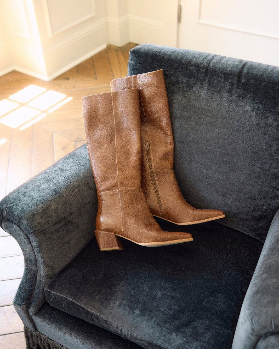 A pair of tan leather knee-high boots with block heels and side zippers, displayed on a dark gray velvet armchair in a room with a wood floor.