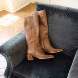 A pair of tan leather knee-high boots with block heels and side zippers, displayed on a dark gray velvet armchair in a room with a wood floor.