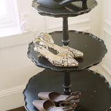 Three black tiered serving trays styled as a shoe stand, holding a variety of women's heels and flats in neutral tones.
