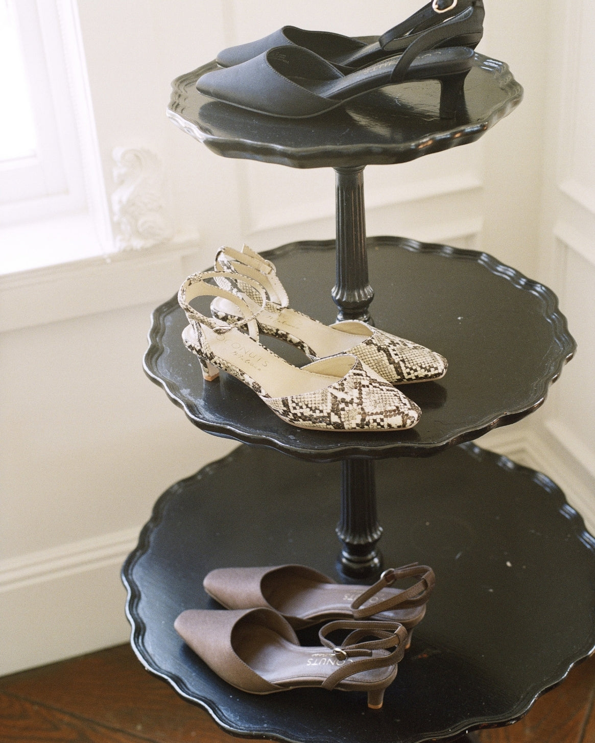 Three black tiered serving trays styled as a shoe stand, holding a variety of women's heels and flats in neutral tones.