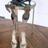 Three pairs of women's slingback heels displayed on a small round metal table and floor: black pointed-toe pumps on the tabletop, taupe slingback mules hanging on the sides, and snakeskin-pattern pointed-toe heels on the floor, set on a wooden floor against a white wall.