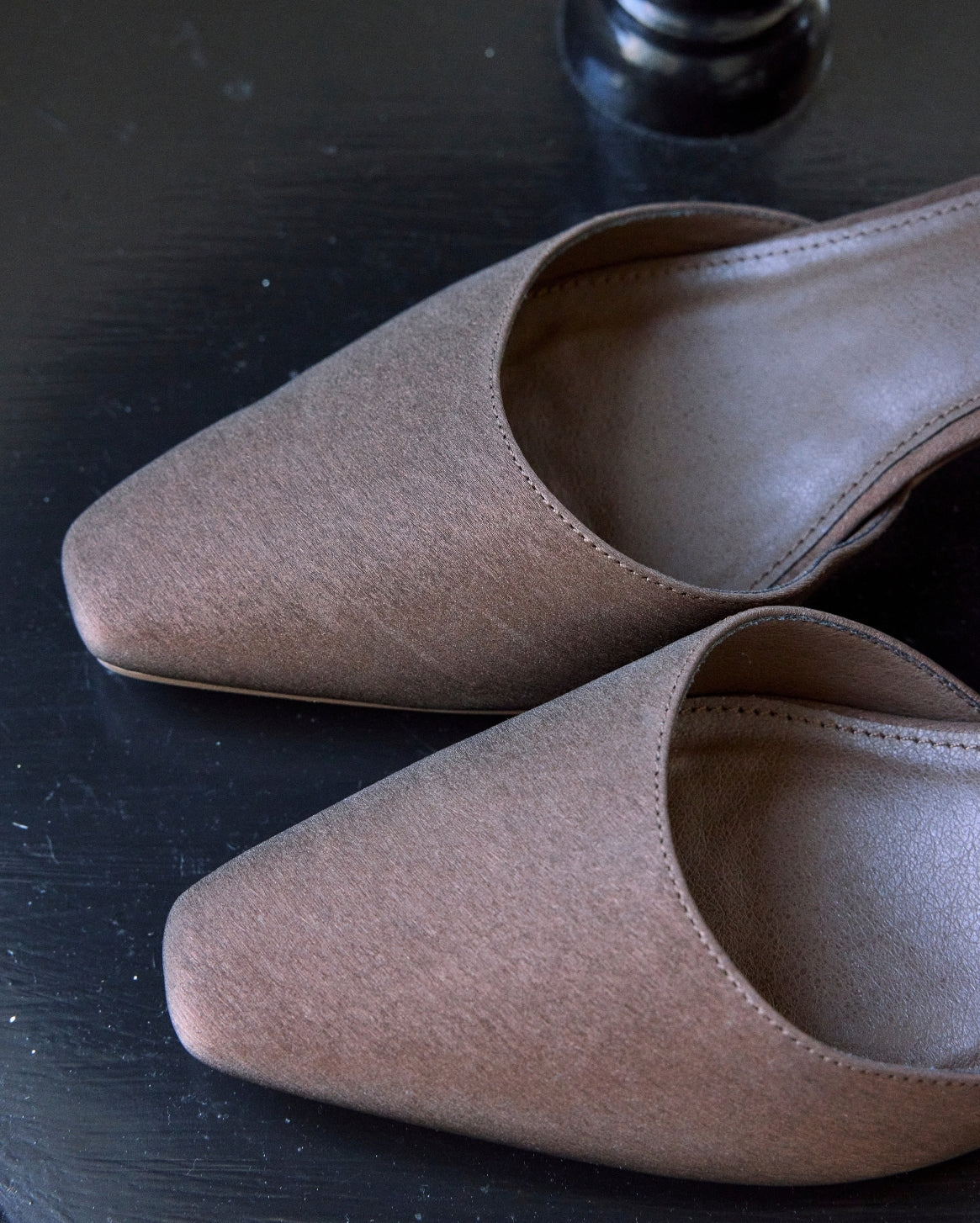 A pair of taupe-brown pointed-toe flat mules with a textured suede-like finish, displayed on a dark wooden surface.