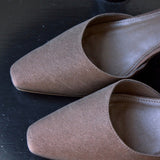 A pair of taupe-brown pointed-toe flat mules with a textured suede-like finish, displayed on a dark wooden surface.