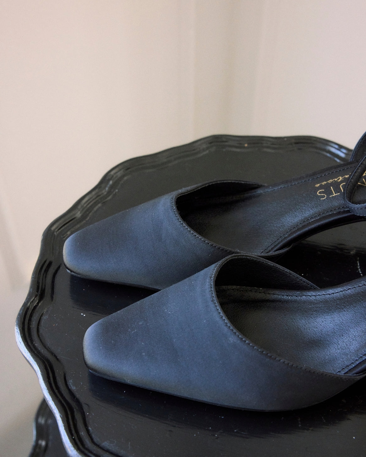A pair of black pointed-toe slingback flats with a satin-like finish, resting on a black scalloped tray against a light beige background.