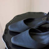 A pair of black pointed-toe slingback flats with a satin-like finish, resting on a black scalloped tray against a light beige background.
