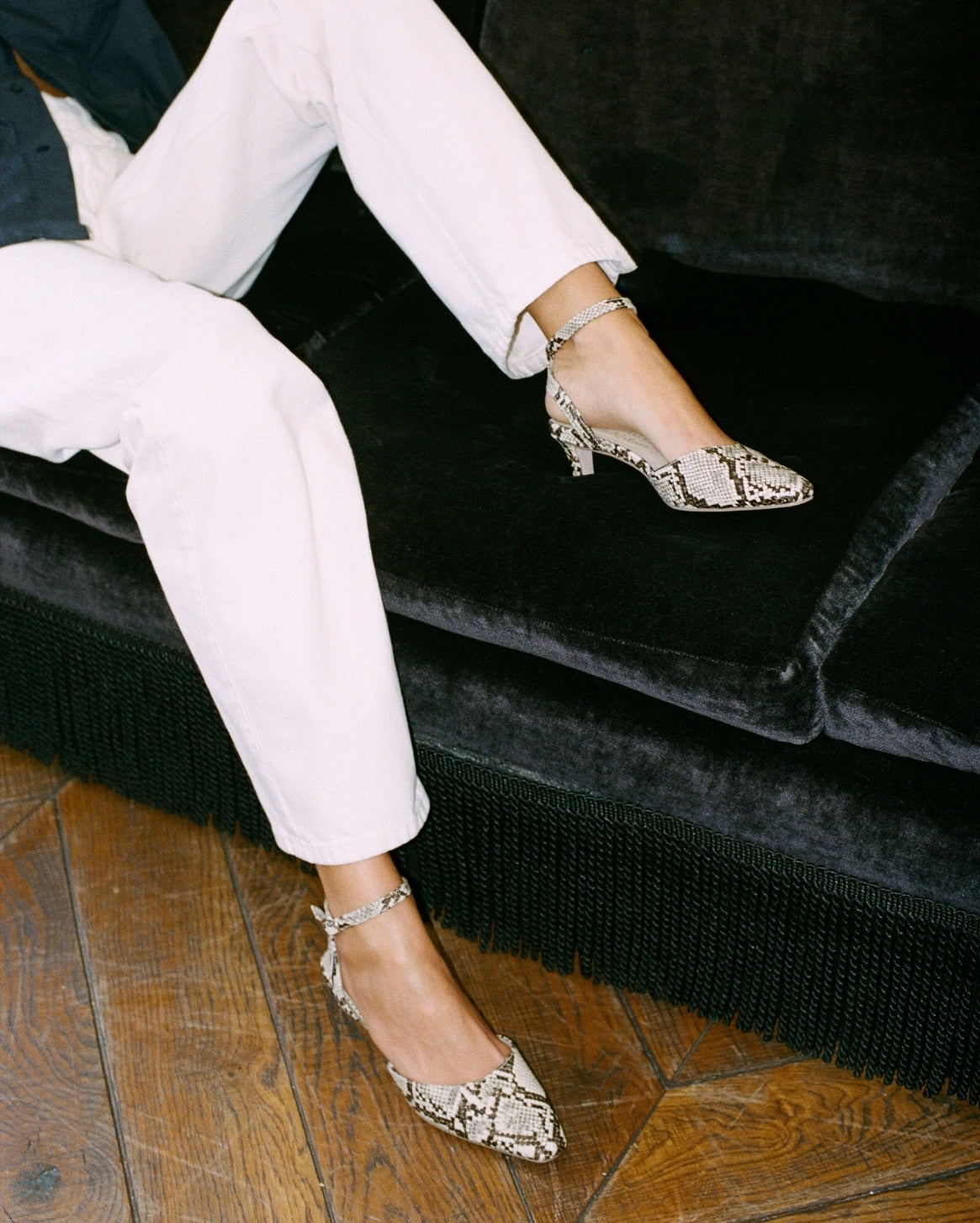 A person seated on a black velvet sofa wearing pointed-toe slingback heels with a snakeskin pattern in beige, brown, and black, paired with white cropped pants and silver ankle straps.
