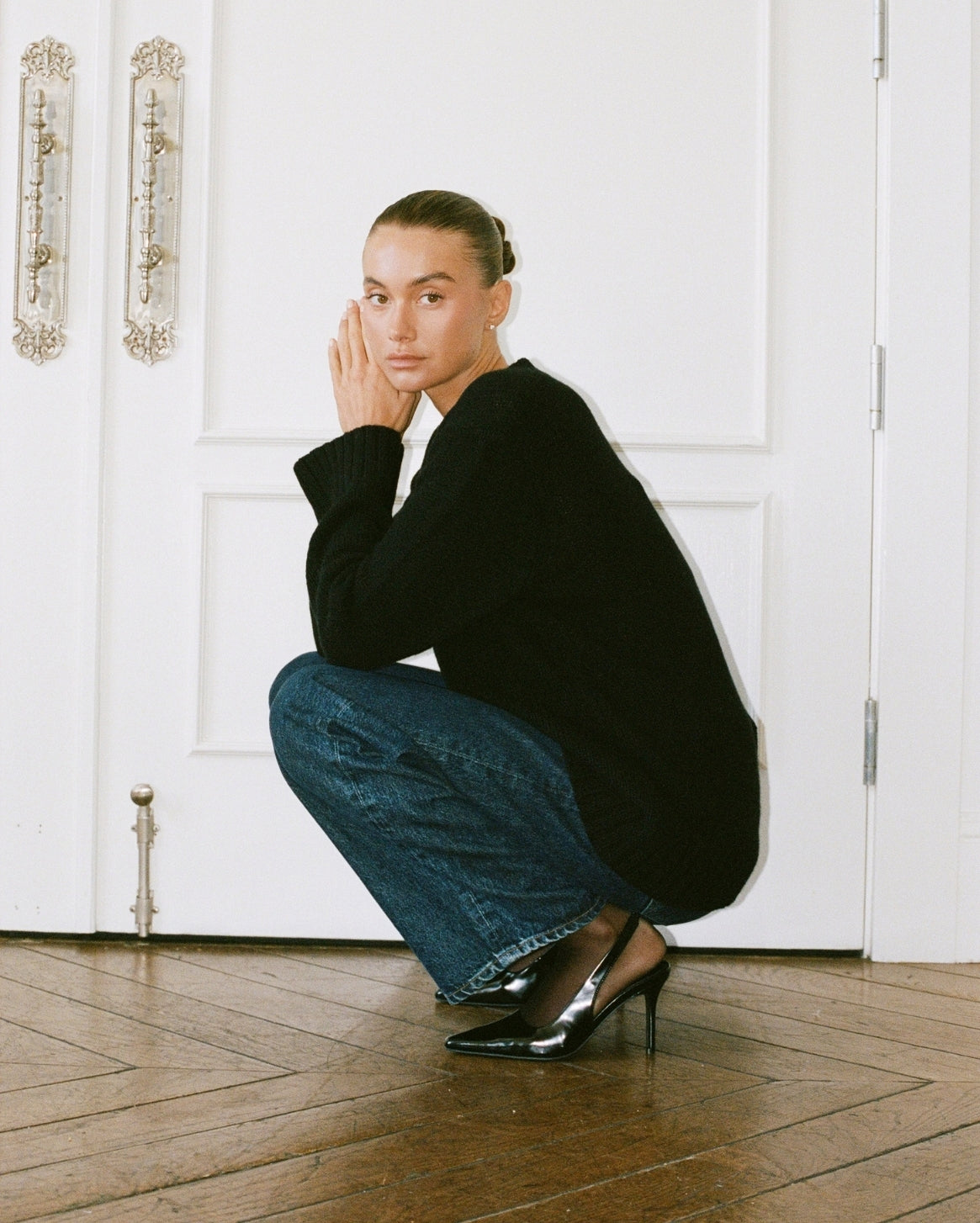 A model crouches on a wooden parquet floor in front of white paneled doors, wearing a black knit cardigan, blue wide-leg jeans, and black pointed-toe heels.