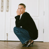A model crouches on a wooden parquet floor in front of white paneled doors, wearing a black knit cardigan, blue wide-leg jeans, and black pointed-toe heels.