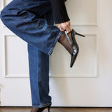 A person wearing blue jeans and a black sweater lifts one leg while holding a black pointed-toe high heel. The other shoe is placed on the wooden floor against a white paneled wall.