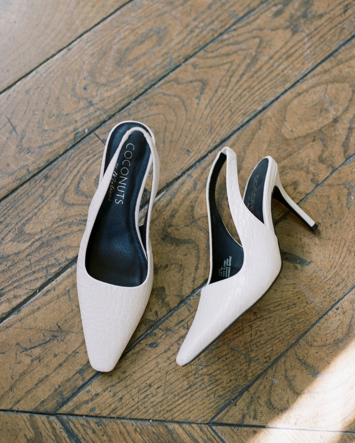 A pair of pointed-toe slingback pumps in a light textured cream color, displayed on a rustic wooden floor. The brand name 'COCONUTS' is visible on the black insoles.