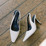 A pair of pointed-toe slingback pumps in a light textured cream color, displayed on a rustic wooden floor. The brand name 'COCONUTS' is visible on the black insoles.
