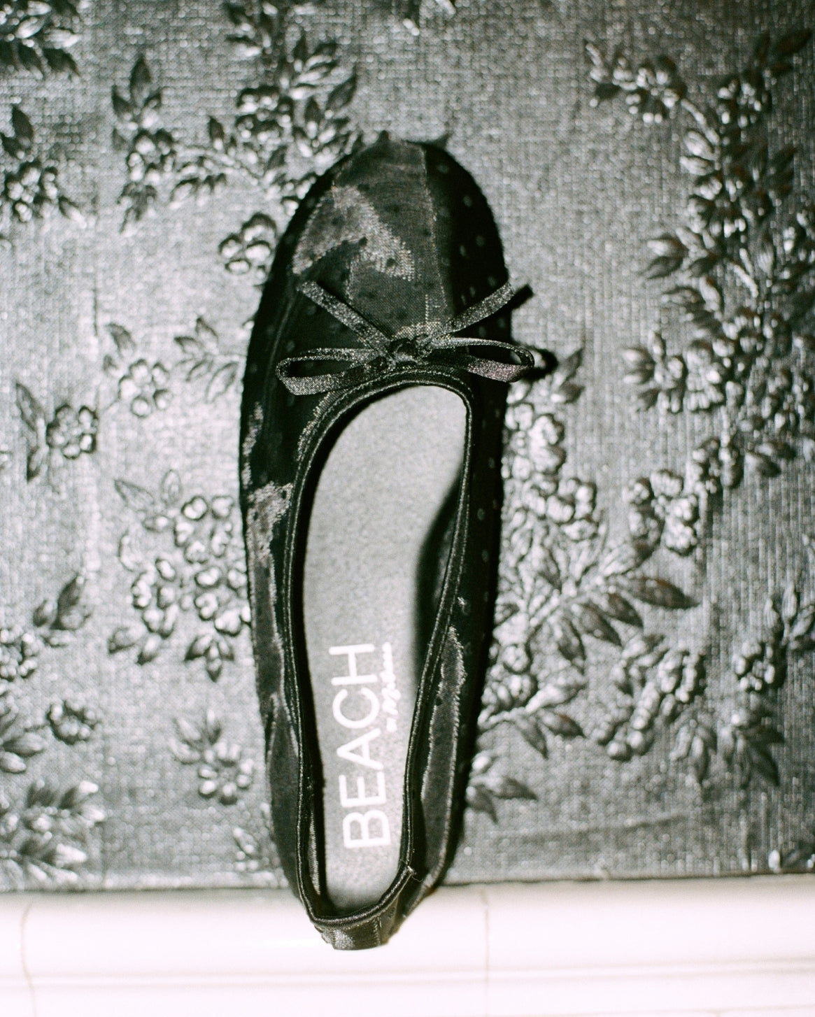 A single black satin ballet flat with a bow on the toe, shown from a top-down view on a textured floral wallpaper background. The insole displays the brand name 'BEACH'.
