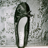 A single black satin ballet flat with a bow on the toe, shown from a top-down view on a textured floral wallpaper background. The insole displays the brand name 'BEACH'.