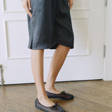 A person wearing a dark charcoal gray midi dress and dark brown patterned ballet flats, standing on a wooden parquet floor in front of a white paneled door.