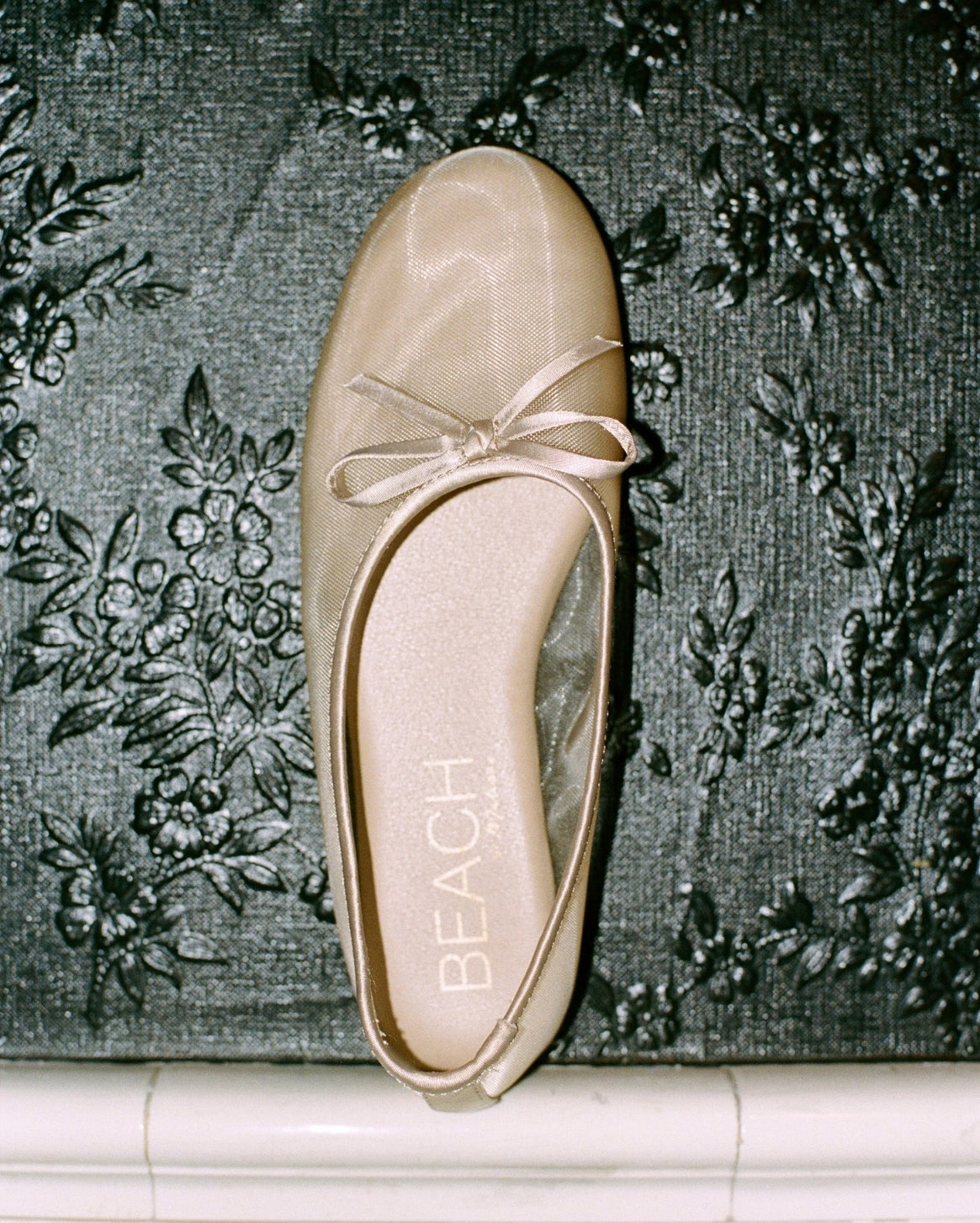 A single champagne-colored satin ballet flat with a bow detail on the toe, resting on a dark floral patterned fabric background. The brand name 'BEACH' is printed on the insole.