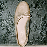A single champagne-colored satin ballet flat with a bow detail on the toe, resting on a dark floral patterned fabric background. The brand name 'BEACH' is printed on the insole.
