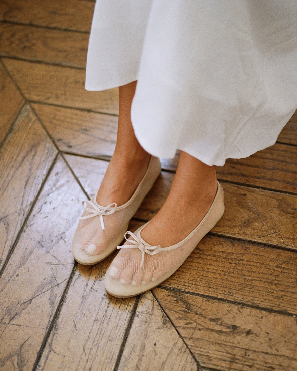 A close-up of a person wearing beige ballet flats with sheer mesh uppers and small bow details, standing on a rustic wooden floor next to a white dress.