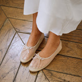 A close-up of a person wearing beige ballet flats with sheer mesh uppers and small bow details, standing on a rustic wooden floor next to a white dress.