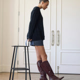 A woman stands in profile indoors wearing a dark navy sweater, denim skirt, and dark burgundy knee-high heeled boots, next to a black metal stool in front of white paneled doors.