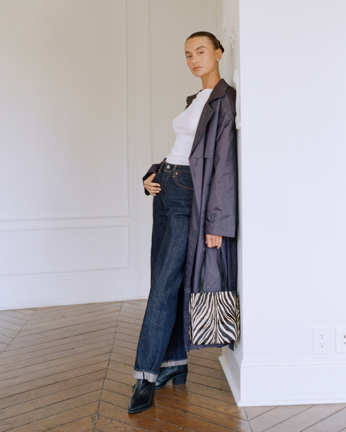 A model stands in a minimalist white room with wood floors, wearing a white top, dark blue wide-leg jeans, black boots, and a dark gray oversized coat, while holding a zebra-print handbag.
