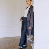 A model stands in a minimalist white room with wood floors, wearing a white top, dark blue wide-leg jeans, black boots, and a dark gray oversized coat, while holding a zebra-print handbag.