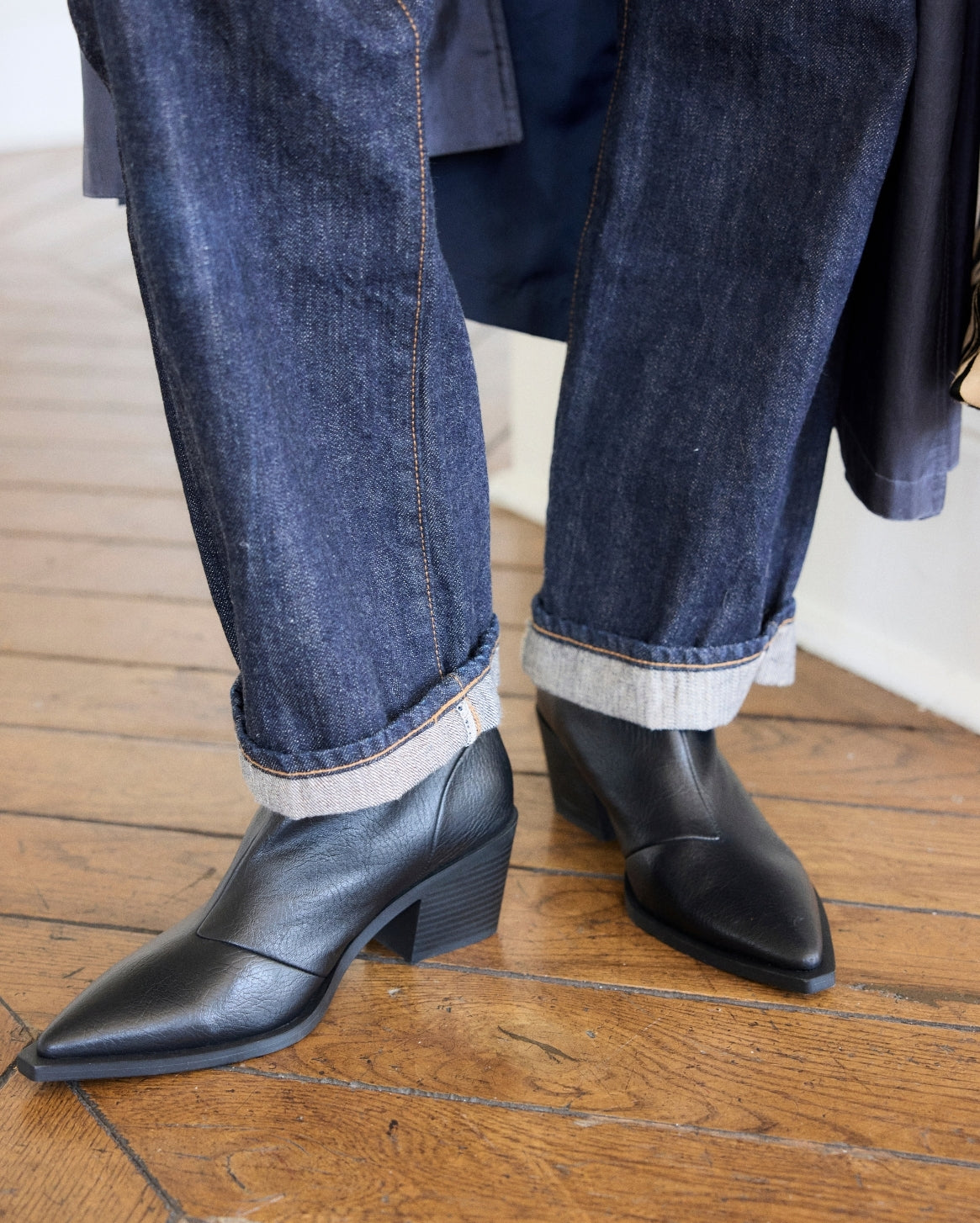 A person wearing dark blue cuffed denim jeans and black pointed-toe ankle boots with low block heels, standing on a wooden floor.