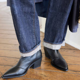 A person wearing dark blue cuffed denim jeans and black pointed-toe ankle boots with low block heels, standing on a wooden floor.