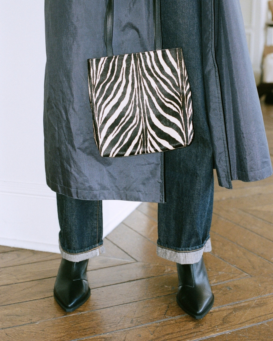 A person wearing a dark gray coat, dark blue jeans with frayed cuffs, and black ankle boots, holding a black and white zebra-print tote bag in a room with a wooden floor.