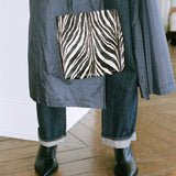 A person wearing a dark gray coat, dark blue jeans with frayed cuffs, and black ankle boots, holding a black and white zebra-print tote bag in a room with a wooden floor.