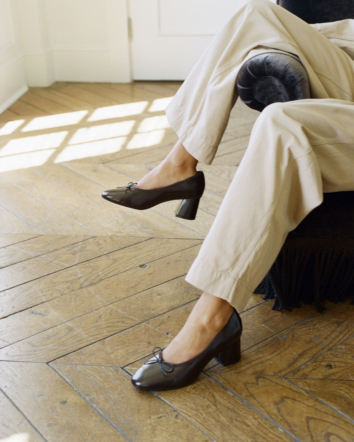 A person seated in a chair on a wooden floor wearing dark brown leather block-heel pumps with small bow details on the toes, paired with light beige trousers.