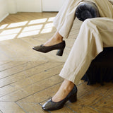 A person seated in a chair on a wooden floor wearing dark brown leather block-heel pumps with small bow details on the toes, paired with light beige trousers.