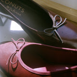 A pair of women's low-heeled flats in a deep burgundy red fabric, featuring a small matching fabric bow on the toe. The insole of the upper shoe displays the brand name 'Coconuts by Matisse'.
