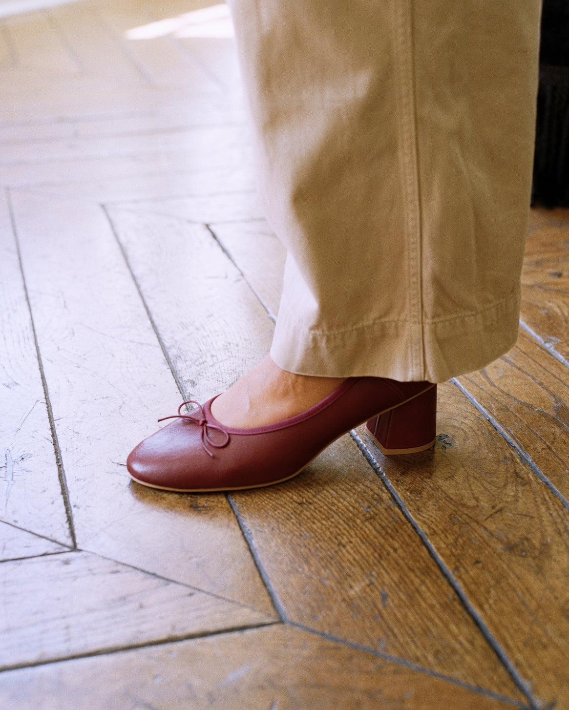 A person wearing beige trousers and a burgundy low-heeled Mary Jane shoe with a small bow on the toe, standing on a wooden parquet floor.