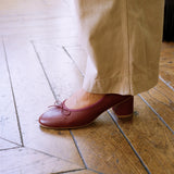 A person wearing beige trousers and a burgundy low-heeled Mary Jane shoe with a small bow on the toe, standing on a wooden parquet floor.