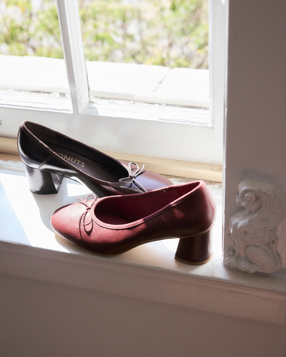 A pair of women's low-heeled pumps with bow details, one in deep burgundy and one in black, displayed on a white window sill.