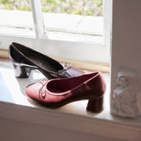 A pair of women's low-heeled pumps with bow details, one in deep burgundy and one in black, displayed on a white window sill.