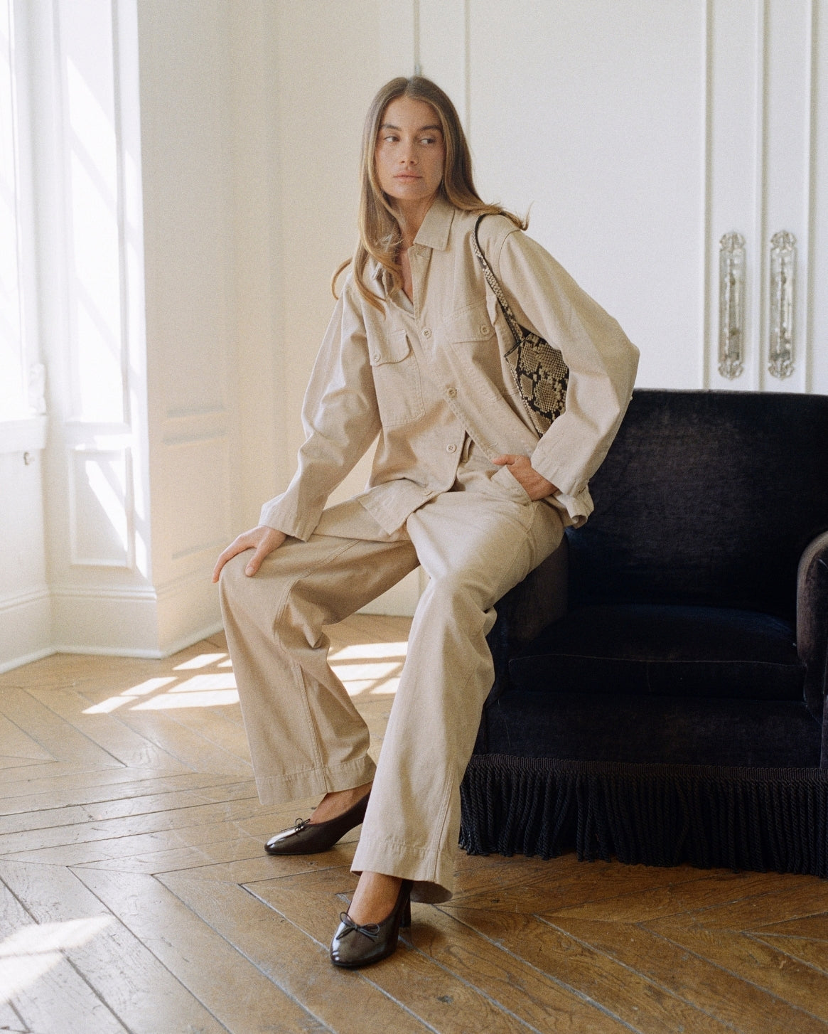 A woman models a beige two-piece linen set consisting of a long-sleeve button-up shirt and matching wide-leg trousers, seated on a dark velvet armchair in a bright room with wooden floors.