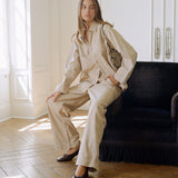 A woman models a beige two-piece linen set consisting of a long-sleeve button-up shirt and matching wide-leg trousers, seated on a dark velvet armchair in a bright room with wooden floors.