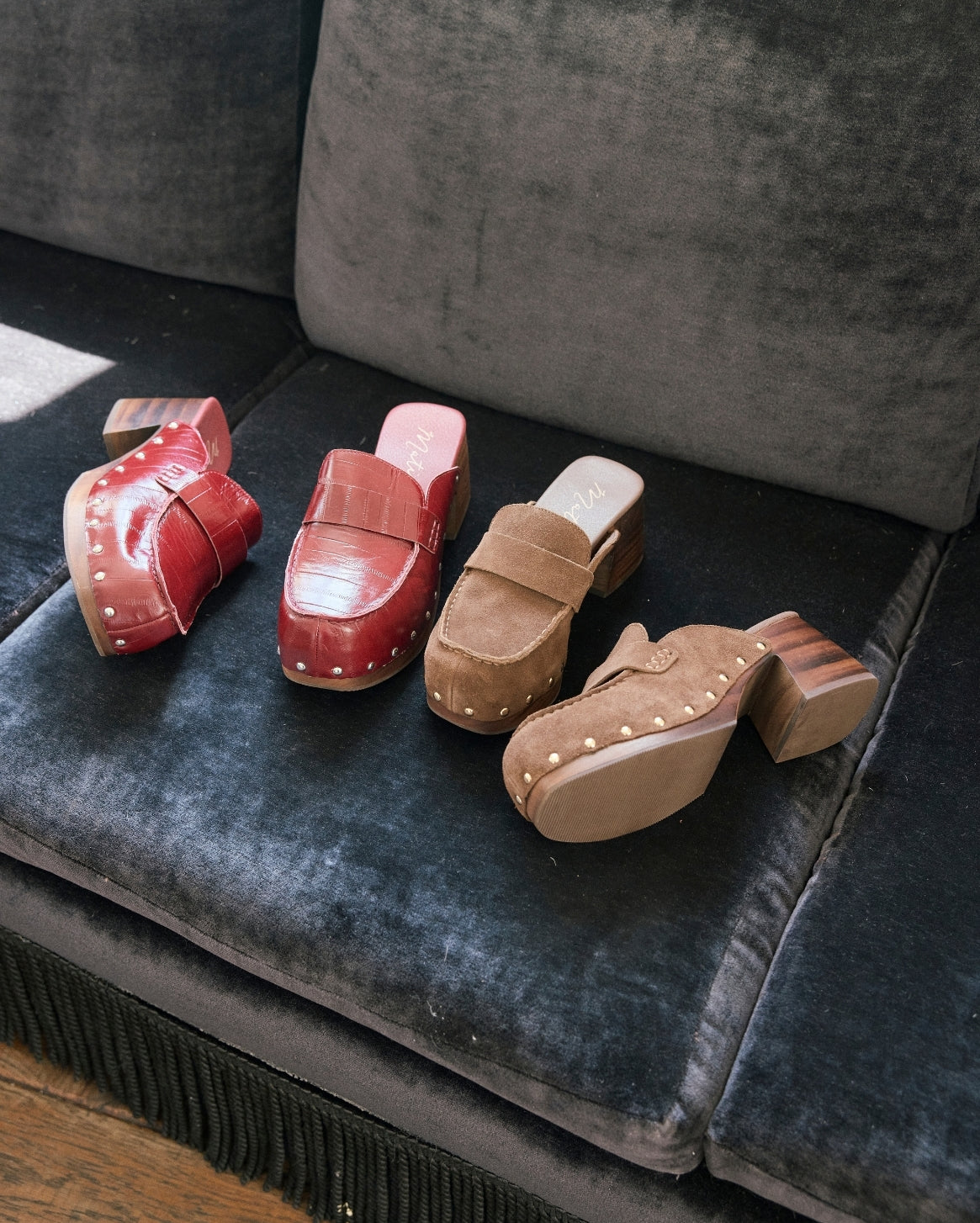 Four clog-style mule shoes in red and brown tones arranged on a dark gray velvet sofa. The shoes have stacked wooden block heels and decorative studs along the sides.