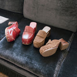 Four clog-style mule shoes in red and brown tones arranged on a dark gray velvet sofa. The shoes have stacked wooden block heels and decorative studs along the sides.