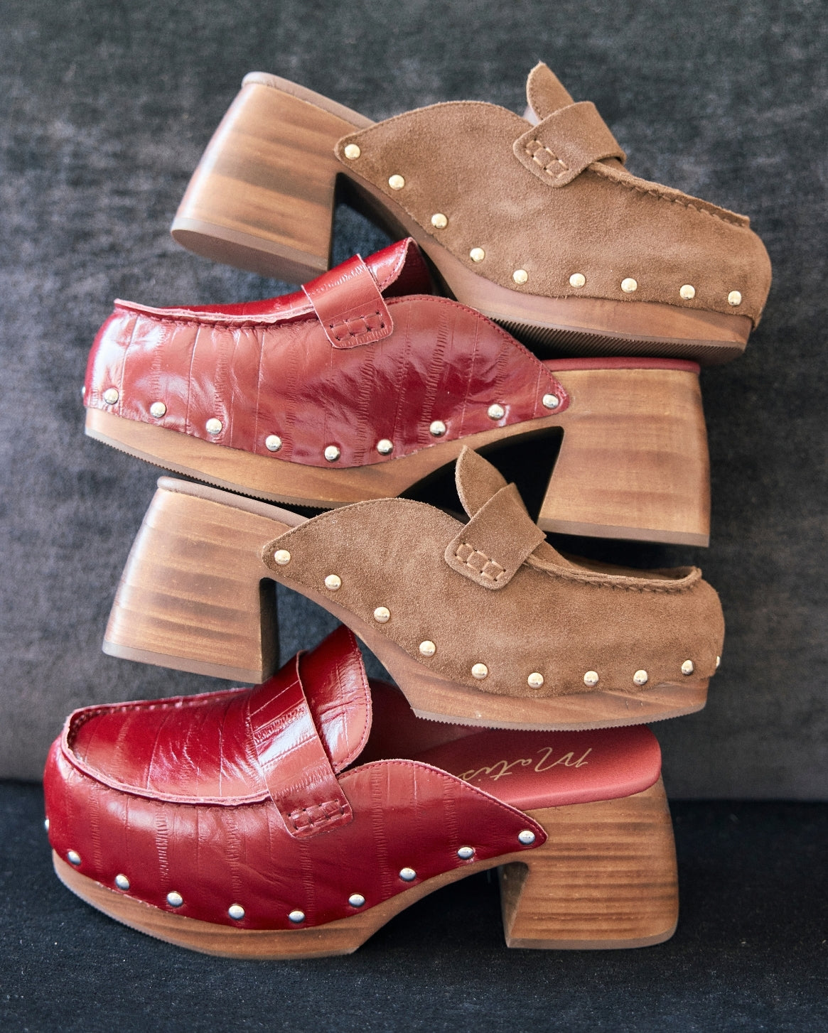 Three pairs of women's clog-style mule shoes stacked vertically on a dark textured background. The top pair is tan suede with small metallic studs and a chunky wooden block heel.