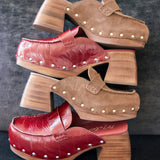 Three pairs of women's clog-style mule shoes stacked vertically on a dark textured background. The top pair is tan suede with small metallic studs and a chunky wooden block heel.