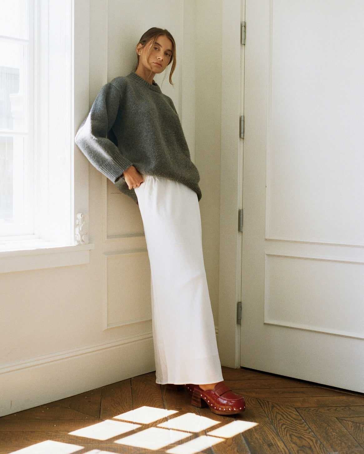 A woman stands in a sunlit room corner wearing a chunky gray knit sweater, a long white skirt, and red clogs.
