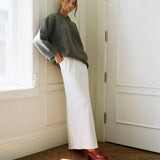 A woman stands in a sunlit room corner wearing a chunky gray knit sweater, a long white skirt, and red clogs.