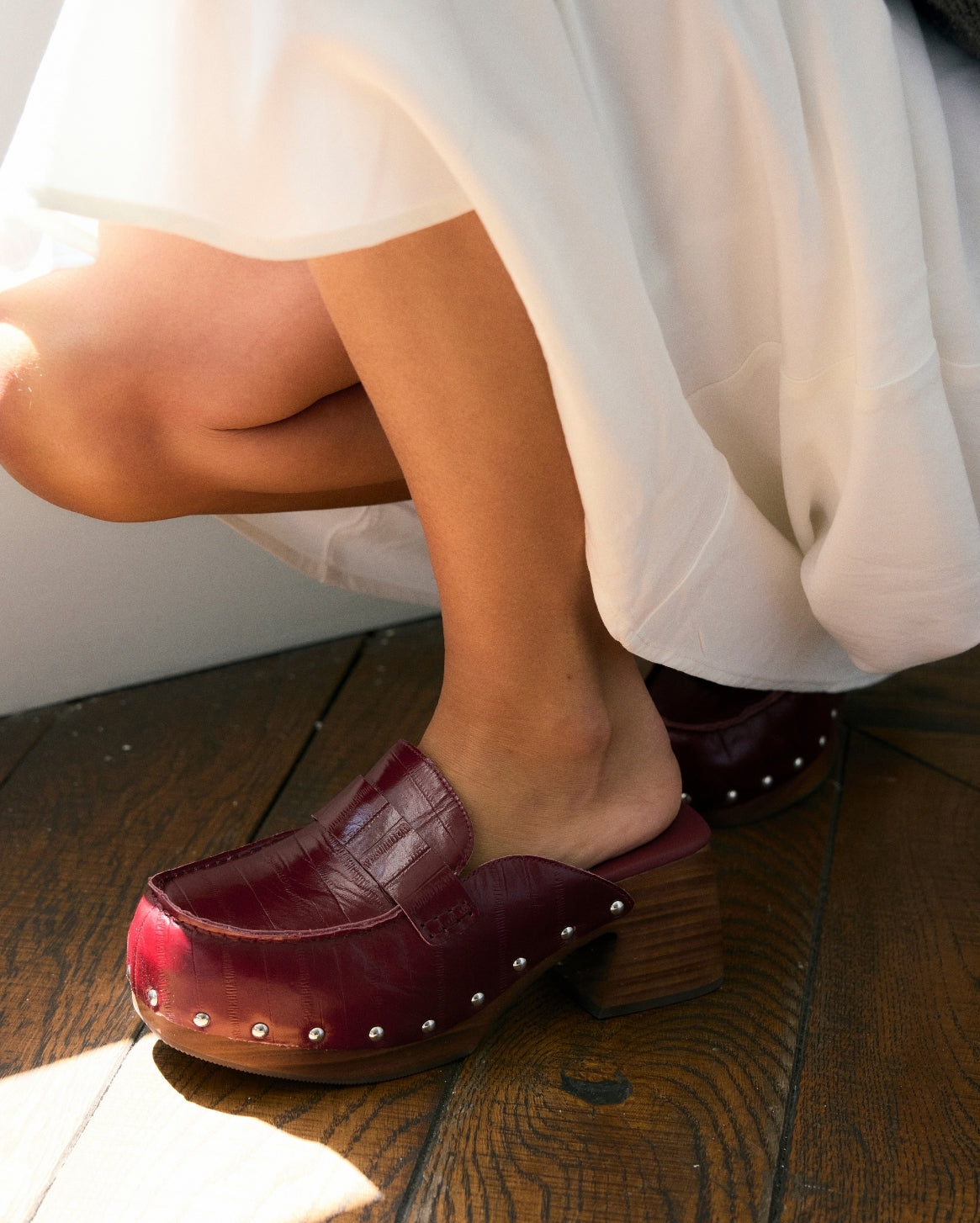 A close-up of a person wearing burgundy leather clog-style mule heels with small silver studs and wooden block heels, seated on a wooden floor with a white dress visible.